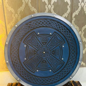 May include: A round, blue shield with a Celtic knot design. The shield has a silver-colored metal rim and is adorned with silver rivets. The intricate black knotwork is centered on the shield's face.