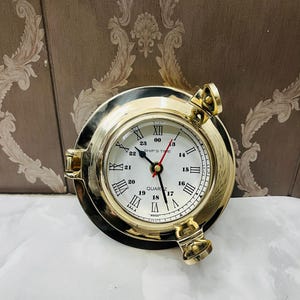 Könnte beinhalten: Eine messingfarbene Quarzuhr im Bullaugen-Design. Das Zifferblatt ist weiß mit schwarzen römischen Ziffern und Zeigern. Die Uhr hat ein maritimes Thema, mit den Worten "SHIP'S TIME" auf dem Zifferblatt.