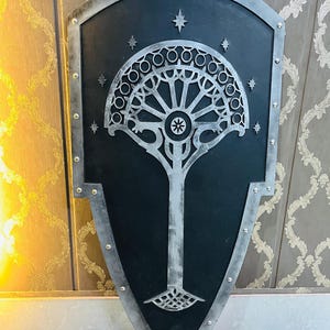 Peut inclure: Un grand objet décoratif en forme de bouclier avec un centre noir et une bordure argentée. Le bouclier présente un motif d'arbre stylisé en argent, avec des étoiles. La bordure est fixée avec des rivets argentés, créant une esthétique médiévale ou fantastique.