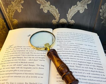 Könnte beinhalten: Eine Lupe mit Holzgriff und Messingrahmen liegt auf einem offenen Buch. Die Seiten des Buches sind mit gedrucktem Text gefüllt. Die Lupe liegt über dem Text und erleichtert das Lesen.