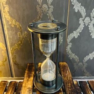 May include: An antique-style hourglass with a dark metal frame and base. The glass bulb contains light-colored sand. The top of the hourglass has a decorative design. The hourglass is set on a wooden surface.
