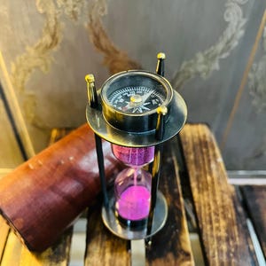 Puede incluir: Un reloj de arena de estilo vintage con una brújula en la parte superior, con un marco de metal negro y arena rosa. El reloj de arena está sobre una superficie de madera, con un estuche de cuero marrón visible. La brújula tiene detalles dorados.
