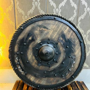 May include: A round wooden shield with a dark metal rim and center boss. The shield features a decorative design and is trimmed with rope. The shield is resting on a wooden slatted surface.