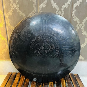 May include: A large, round, black metal decorative tray with intricate etched patterns. The tray is resting on a wooden slatted base. The background features a patterned wallpaper.