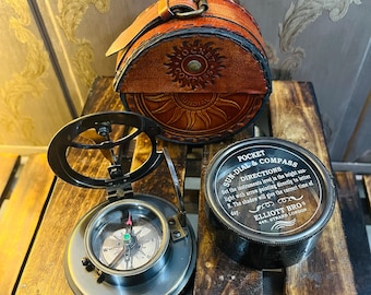 Puede incluir: Un juego de brújula y reloj de sol vintage con una funda de cuero marrón. La brújula está abierta, mostrando la aguja y las marcas direccionales. La funda de cuero presenta un diseño de sol. También hay un pequeño recipiente negro con texto.