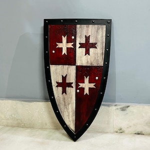 May include: A medieval-style shield with a black border and rivets. The shield is divided into four quadrants, alternating between red and white, each featuring a white cross design. The shield has a distressed, aged appearance.
