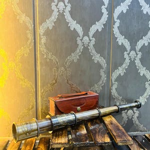 May include: A vintage brass telescope and a brown leather case. The telescope is lying on a wooden stand. The case has a handle and a clasp. The telescope is approximately 30 cm long.