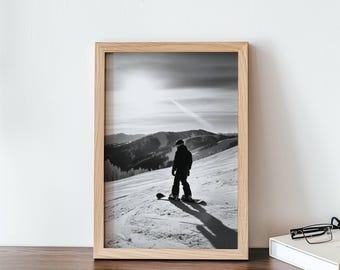 Puede incluir: Fotografía en blanco y negro de una persona haciendo snowboard por una ladera de montaña nevada. La persona lleva una chaqueta y un gorro. El sol brilla intensamente en el fondo.