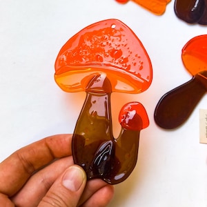 May include: A glass mushroom ornament with a red and orange cap and a brown stem. The mushroom is hand-blown and has a unique, organic shape.