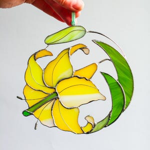 Op de afbeelding: Een gebrandschilderd zonnenvanger met een gele lelie en groene bladeren. De lelie bevindt zich in het midden van de zonnenvanger en is omringd door groene bladeren. De zonnenvanger is gemaakt van glas en wordt vastgehouden door een hand.