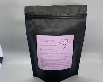 Op de afbeelding: Een zwarte zak van Magnesium Bath Ritual, met een roze label met tekst en een bloemenillustratie. De tekst luidt "Sleep & Let Go" en "I have done enough for today, I choose sleep, rest & peace." Een decoratieve bloem ligt ervoor.