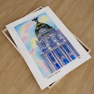 May include: Watercolor painting of a building in Madrid, Spain. The building is a tall, narrow structure with a large dome on top. The dome is decorated with a golden statue of a winged figure. The building is painted in shades of purple, blue, and white. The sky is a light blue with pink and yellow clouds.
