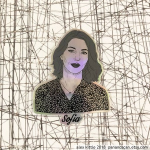 May include: Holographic sticker featuring a portrait of a woman with short, wavy hair and a patterned top. The sticker has the name "Sofia" written in cursive. The background is a complex, geometric pattern.