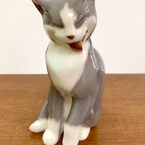 May include: A glossy porcelain figurine of a seated cat with a smooth, grey and white coat. The cat is depicted in a grooming pose with its head tilted, one paw raised, and its tongue extended. The finish is reflective with soft color transitions.