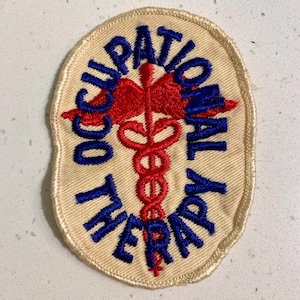 May include: An embroidered patch with the words "OCCUPATIONAL THERAPY" in blue, arched around a red caduceus symbol. The patch has a cream-colored background and a stitched border.