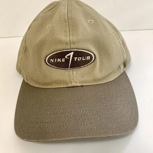 May include: A tan baseball cap with a structured crown and a darker brown brim. The front features an oval-shaped patch with a brown background and white text that reads NIKE TOUR, with a stylized white swoosh logo integrated into the design.