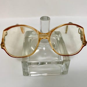 May include: A pair of amber-colored plastic eyeglasses with round lenses. The glasses are sitting on a clear glass display stand.