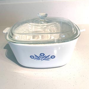 May include: A white square casserole dish with rounded corners and integrated side handles. The front features a blue floral pattern. It includes a clear, square glass lid with a central knob handle, providing a view into the interior.