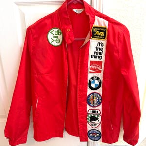 May include: A bright red zip-up jacket featuring a white vertical stripe on the right side adorned with various patches including BMW, Alfa Romeo, and Coca-Cola logos, plus a circular green patch on the left chest with the text db and symbols.