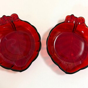 May include: Two matching translucent red glass dishes shaped like leaves. Each dish features a scalloped edge, a central vein pattern embossed on the interior surface, and a decorative stem-like handle at the top with a curled design element.