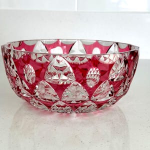 May include: A round crystal bowl featuring a deep cranberry red color overlaid on clear glass. The exterior surface is intricately cut with a repeating pattern of geometric triangles and oval shapes, creating a faceted texture throughout the piece.
