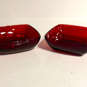 May include: Two small, square-shaped bowls crafted from translucent, deep red glass. Each bowl features rounded corners, smooth curved sides, and a wide, open top, presenting a consistent, uniform design across both individual glass pieces.