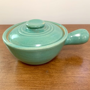 May include: A teal ceramic soup bowl with a lid and a handle. The bowl has a round shape with a lid that fits snugly on top. The handle is curved and easy to grip. The bowl is made of ceramic and has a smooth, glossy finish.