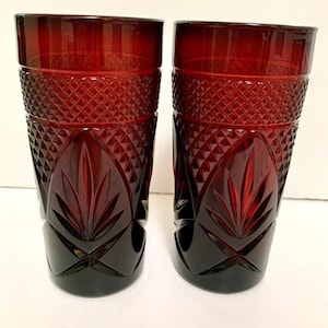 May include: Two tall, cylindrical drinking glasses made of deep red glass. Each glass features a textured diamond pattern around the upper section and a prominent, embossed leaf or petal design around the lower half, creating a faceted appearance.