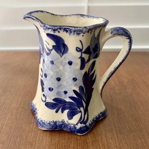 May include: A cream-colored ceramic pitcher featuring a hand-painted blue floral pattern with hydrangea-like clusters and leafy stems. The pitcher has a scalloped rim and base, a curved handle, and a blue-sponged decorative edge along the top and bottom.