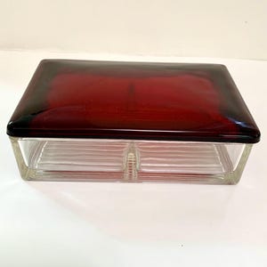 May include: A rectangular glass box featuring a deep red, smooth, slightly domed lid and a clear, transparent base. The base has a ribbed, horizontal texture on the interior walls and a central vertical divider, creating two distinct storage sections.