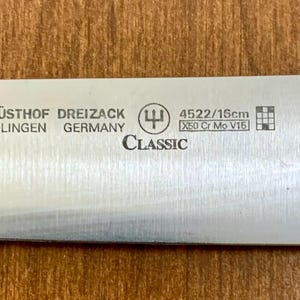 May include: Close-up of a stainless steel knife blade with the text "W&Uuml;STHOF DREIZACK SOLINGEN GERMANY CLASSIC". The blade has the markings "4522/16cm" and "X50 Cr Mo V15". The knife is 16cm long.