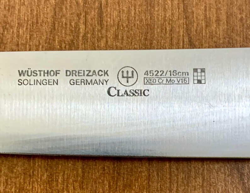 May include: Close-up of a stainless steel knife blade with the text "W&Uuml;STHOF DREIZACK SOLINGEN GERMANY CLASSIC". The blade has the markings "4522/16cm" and "X50 Cr Mo V15". The knife is 16cm long.
