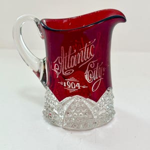 May include: A ruby red glass pitcher with a clear glass handle and a clear, diamond-patterned base. The front features white etched text that reads Atlantic City and 1904. The pitcher has a curved spout and a smooth, translucent upper body.