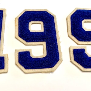 May include: Three individual iron-on patches featuring the numbers 1, 9, and 9. Each patch has a vibrant blue textured center with a thick, off-white felt border. The numbers are designed in a classic block font style with sharp, angular edges.