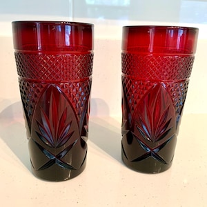 May include: Two ruby red, cut-glass tumblers. The glasses feature a diamond pattern near the top, with a leaf-like design at the base. The bottom portion of the glasses is a darker shade of red.