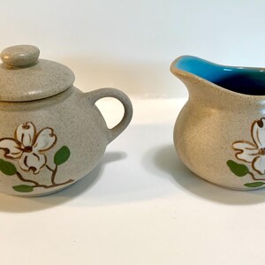 May include: A vintage ceramic sugar and creamer set with a floral design. The creamer has a light blue interior and the sugar bowl has a lid. Both pieces feature a white flower with brown stems and green leaves.
