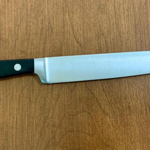 May include: A stainless steel chef's knife with a black handle. The blade is silver and tapers to a sharp point. The handle has three silver rivets. The knife is lying on a wooden surface.