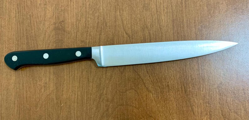May include: A stainless steel chef's knife with a black handle. The blade is silver and tapers to a sharp point. The handle has three silver rivets. The knife is lying on a wooden surface.