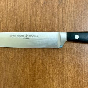 May include: A stainless steel chef's knife with a black handle. The blade is long and slender, with the text "W&Uuml;STHOF DREIZACK SOLINGEN GERMANY CLASSIC" printed on the blade. The handle has three white rivets.