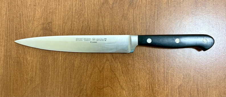 May include: A stainless steel chef's knife with a black handle. The blade is long and slender, with the text "W&Uuml;STHOF DREIZACK SOLINGEN GERMANY CLASSIC" printed on the blade. The handle has three white rivets.
