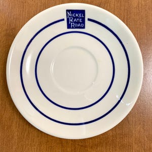 May include: A white ceramic saucer featuring two concentric dark blue circular lines and a dark blue square logo at the top containing the text Nickel Plate Road in white lettering. The saucer has a central circular indentation for a cup.