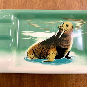 May include: Rectangular ceramic ashtray with a green and white gradient background. A detailed illustration of a walrus with tusks is centered on the tray. Gold accents and handwritten text are visible.