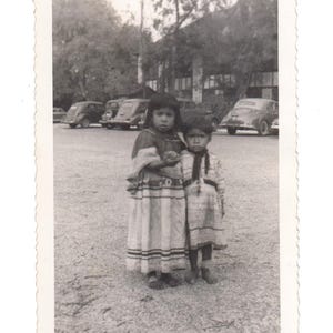 May include: Black and white photo of two young girls wearing traditional clothing standing in front of a building. The girls are smiling and looking at the camera.