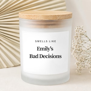 Puede incluir: Una vela en frasco de vidrio esmerilado con tapa de madera clara. La etiqueta rectangular blanca en el frente presenta el texto negro "SMELLS LIKE" sobre el texto negro más grande "Emily's Bad Decisions" en una fuente con remates.