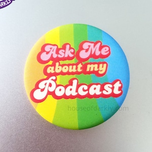 May include: A rainbow-coloured button with the text "Ask Me about my Podcast" in pink and white letters with a retro font. The text "houseofdarkly.com" is printed in black at the bottom of the button.