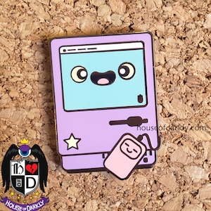 Peut inclure: Une épingle en émail de couleur violette et bleue représentant un ordinateur de dessin animé avec un téléphone rose attaché. L'ordinateur a un visage joyeux et une étoile sur le côté. Le téléphone a également un visage joyeux. L'épingle est sur un fond de tableau en liège.