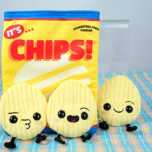 May include: Three plush chip-shaped toys with smiling faces. The chips are yellow and have a textured surface. The chips are in front of a yellow bag with the words "It's... Chips!" printed on it.