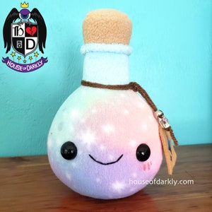 May include: A plush toy shaped like a potion bottle with a cork stopper. The bottle is a rainbow gradient of pink, purple, blue, and green with white stars. It has a brown string tied around the neck and a small tag with the text "houseofdarkly.com".