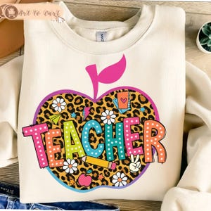 Op de afbeelding: Crèmekleurige sweatshirt met een kleurrijk appelontwerp. De appel heeft een luipaardprintpatroon, madeliefjes en het woord "TEACHER" in heldere, gestippelde letters. Extra afbeeldingen zijn een potlood, vredesteken en een kleine appel.