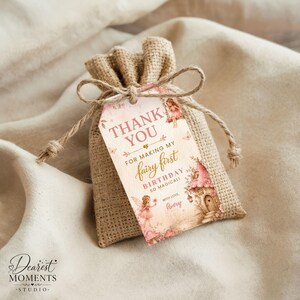 May include: A tan hessian drawstring favour bag with a rectangular paper tag attached by twine. The tag features pink fairy illustrations, a mushroom house, and text reading: THANK YOU FOR MAKING MY fairy first BIRTHDAY SO MAGICAL! WITH LOVE, Avery.